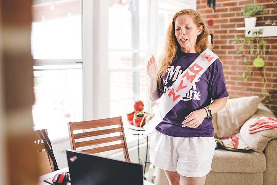 Teacher in her home, wear Muse t-shirt and "womyn" sash, standing and physically interacting, in front of computer