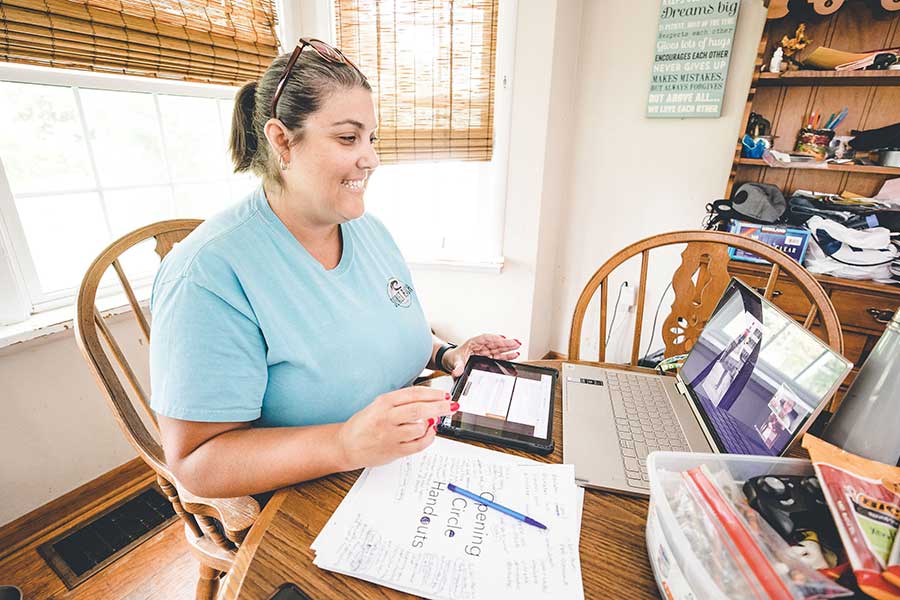 Smiling teacher in her home, at a table with art supplies, notes and a computer, with virtual Institute in progress