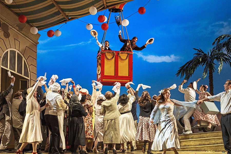 Image from The Elixir of Love opera with a small crowd of people dressed in white waving to two people floating away in the basket of a hot air balloon.