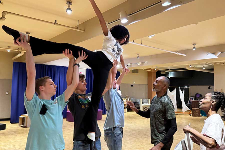 Three student performers lift a dancer above their heads, instructed by two choreographers